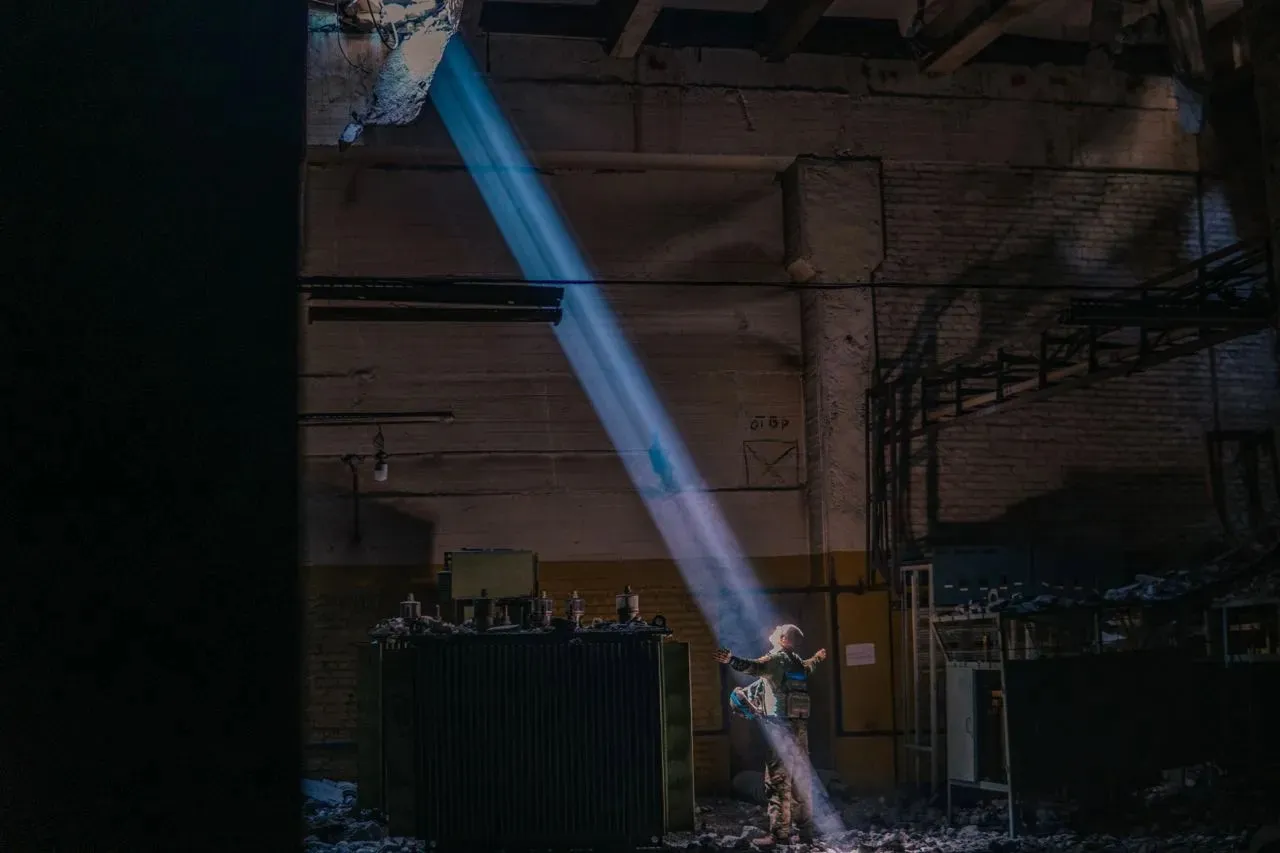 A beam of light falls through the shattered roof of a workshop into the darkness, onto a soldier standing with arms spread wide